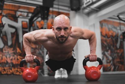 Man doing advanced push-ups with hands placed on two red kettlebells for added range of motion.